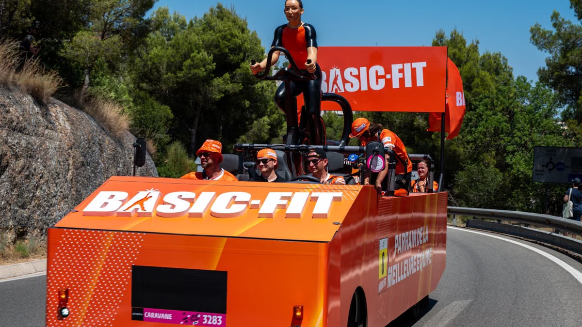 Colorful Basic-Fit promotional vehicle driving on a scenic road during a sunny day parade.